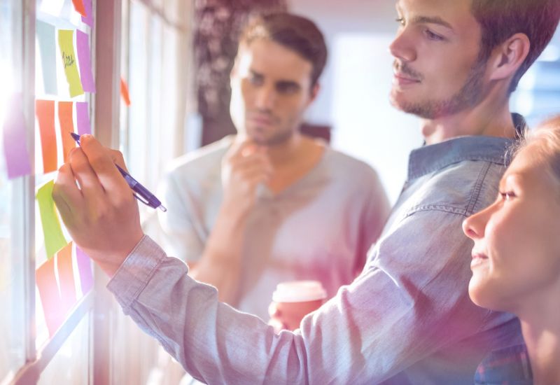 person writing on a sticky note with 2 other watching