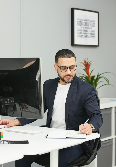 man at an office desk behind a computer, leaning to write on a note pad